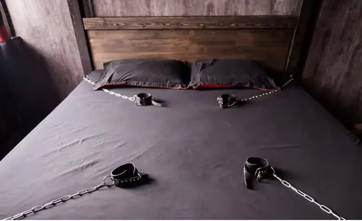 a bad in a dimly lit room with restraints attached to a bed with grey sheets